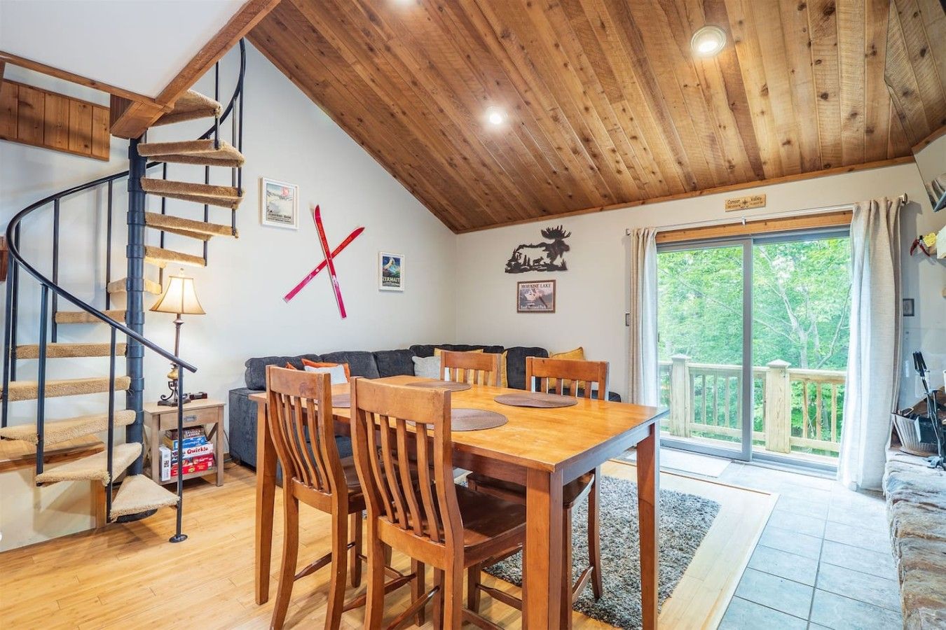 A cozy interior with a wooden staircase, a dining area with wooden chairs, a sofa, and a large window offering a view of greenery.