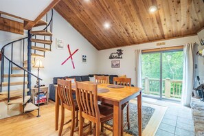 A cozy interior with a wooden staircase, a dining area with wooden chairs, a sofa, and a large window offering a view of greenery.