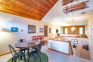 A room with a wooden ceiling, a round table with chairs, a bar area with a counter, and various framed pictures on the walls.