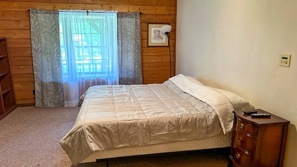 A bedroom with a bed, a nightstand, a lamp, a window with curtains, and a thermostat on the wall. - Back Country Cabin - Stunning, Secluded Property (Davis)