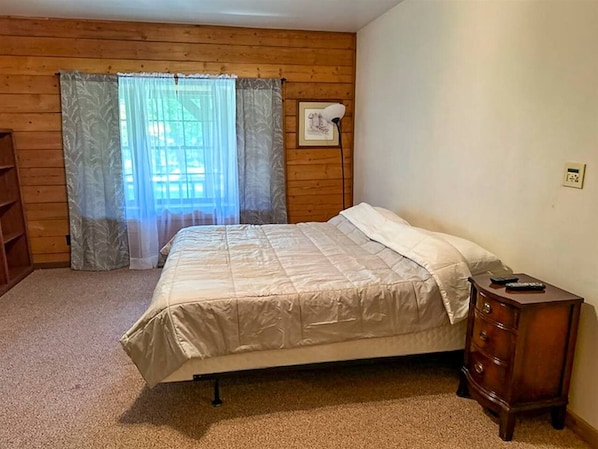 A bedroom with a bed, a nightstand, a lamp, a window with curtains, and a thermostat on the wall.