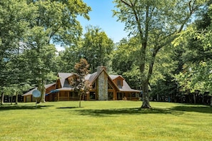 A log cabin with a stone chimney, surrounded by a well-maintained lawn and dense trees.