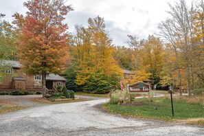 A gravel driveway leading to a wooden sign with 'Woods End' written on it, surrounded by autumn-colored trees.