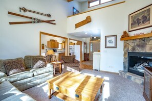 A spacious living room with a fireplace, a kitchen in the background, and ski equipment on the wall.