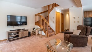 A modern living room with a flat-screen TV, wooden entertainment unit, glass coffee table, brown leather sofa, and a wooden staircase. - Deerfield Village 099 (Davis)