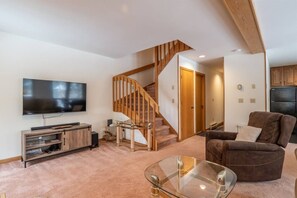 Un salon moderne doté d’un téléviseur à écran plat, d’une table basse en verre, d’un canapé en cuir marron, d’un escalier en bois et d’un coin cuisine avec des meubles en bois.