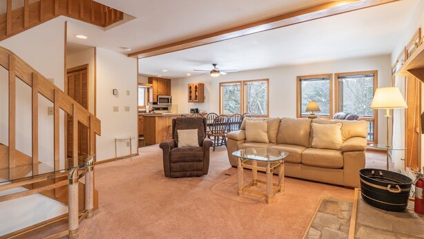 A spacious living area with a sofa, armchair, and glass coffee table. A kitchen is visible in the background, and a staircase leads to an upper level. - Deerfield Village 099 (Davis)