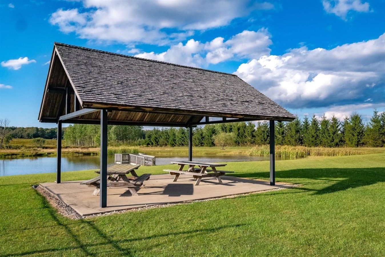 A covered picnic area with benches and picnic tables, surrounded by a grassy field and a body of water.