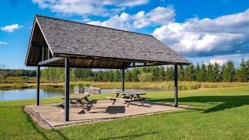 A covered picnic area with benches and picnic tables, surrounded by a grassy field and a body of water.