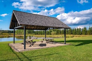 A covered picnic area with benches and picnic tables, surrounded by a grassy field and a body of water.
