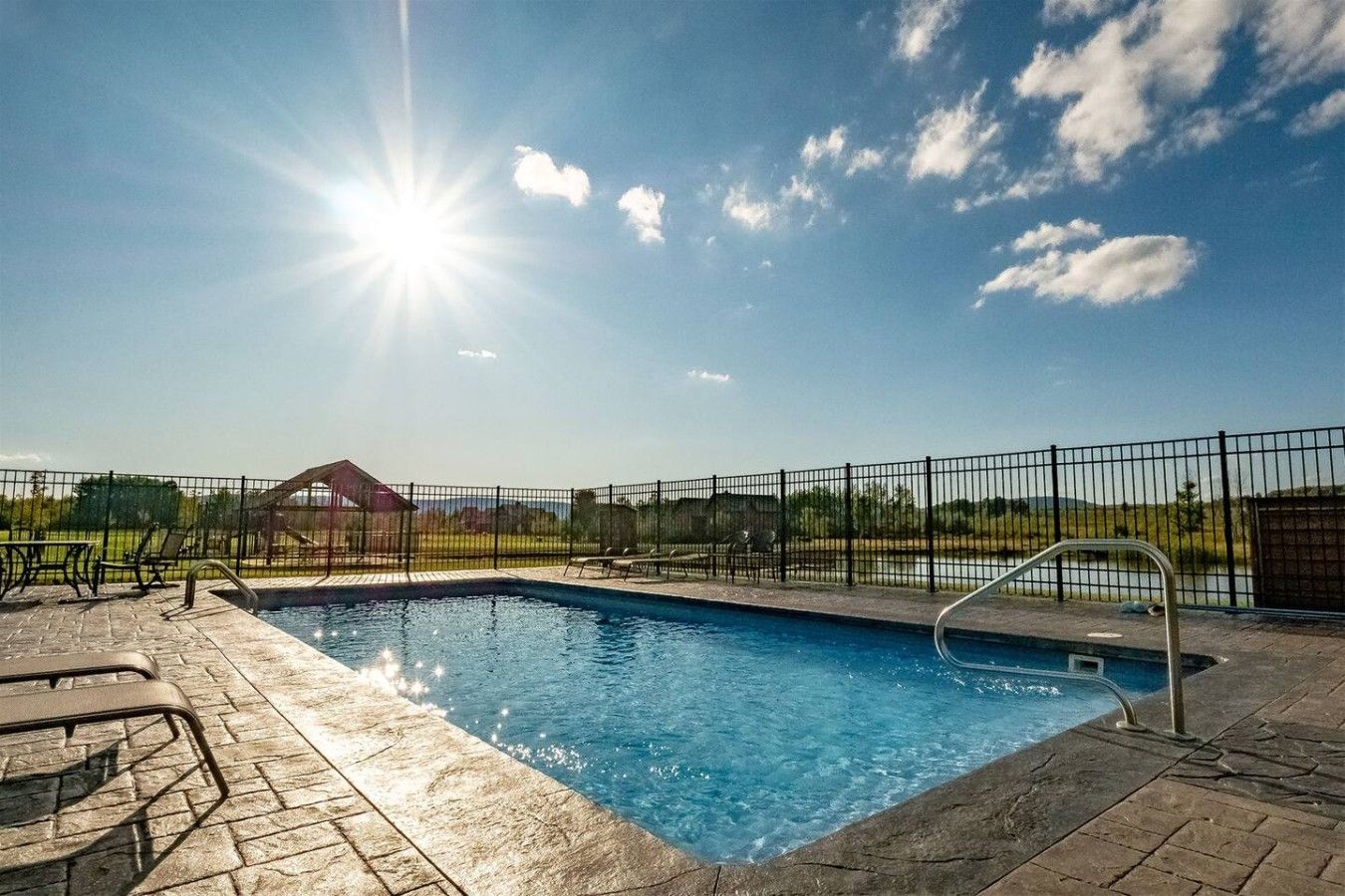 A swimming pool with a metal ladder, surrounded by a black fence, and a clear sky with the sun shining.