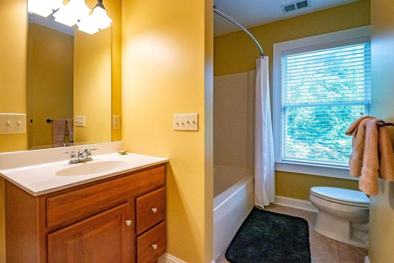Une salle de bain avec un lavabo blanc, un meuble-lavabo en bois, un rideau de douche et une fenêtre avec des stores.
