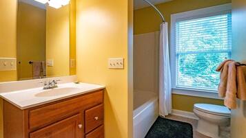 Une salle de bain avec un lavabo blanc, un meuble-lavabo en bois, un rideau de douche et une fenêtre avec des stores.