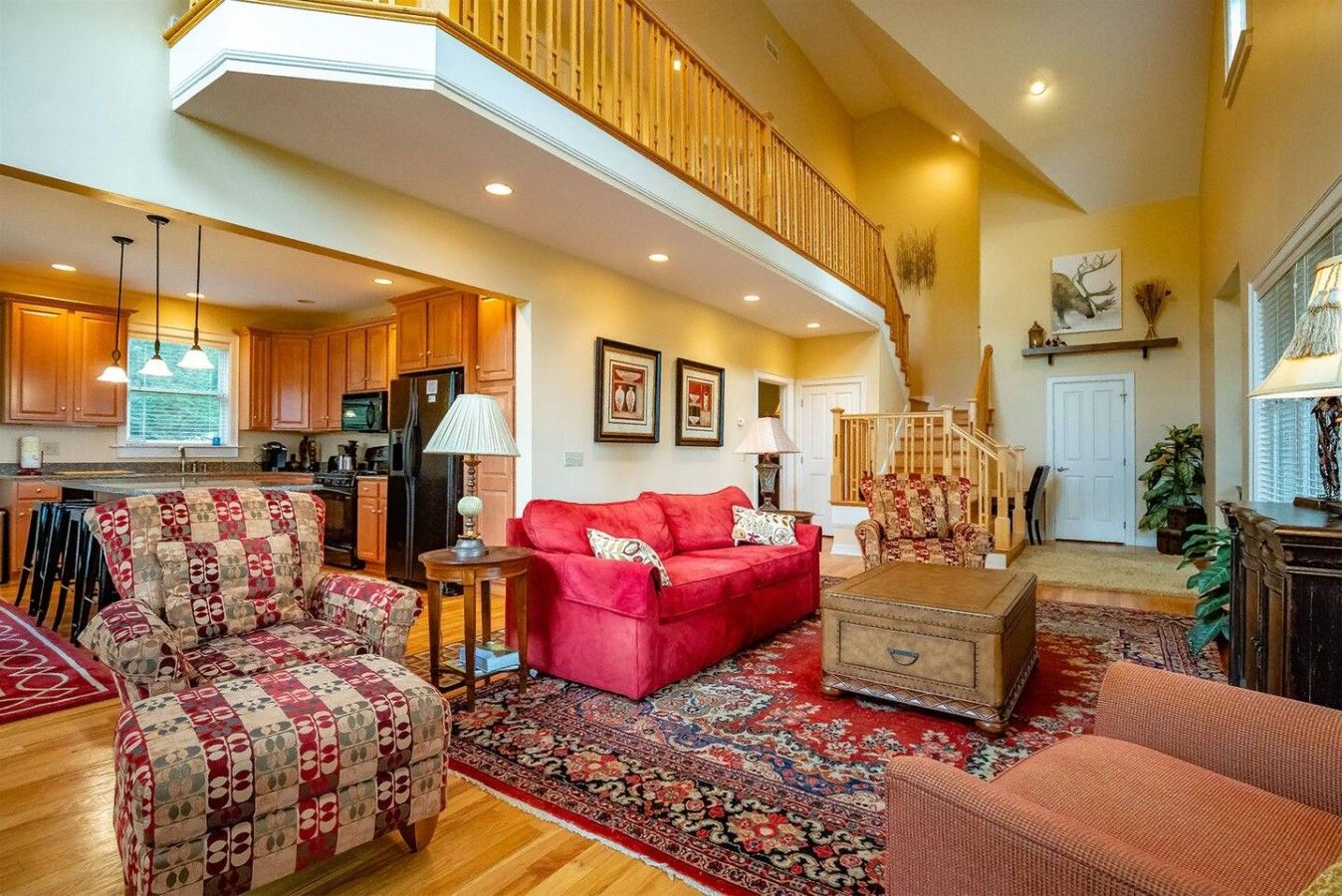A spacious living room with a red sofa, a patterned armchair, and a coffee table. A kitchen is visible in the background with wooden cabinets and a refrigerator.