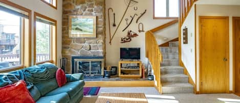 A living room with a stone fireplace, a television, a staircase, and a large window.