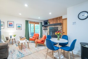 A modern living room with a fireplace, a dining area with blue chairs, and a large window with curtains.