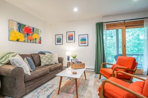 A living room with a brown sofa, a white coffee table, and an orange chair. There are framed pictures on the wall and a window with curtains.