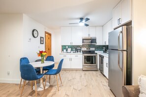 A modern kitchen with white cabinets, stainless steel appliances, a dining area with blue chairs, and a wooden floor.