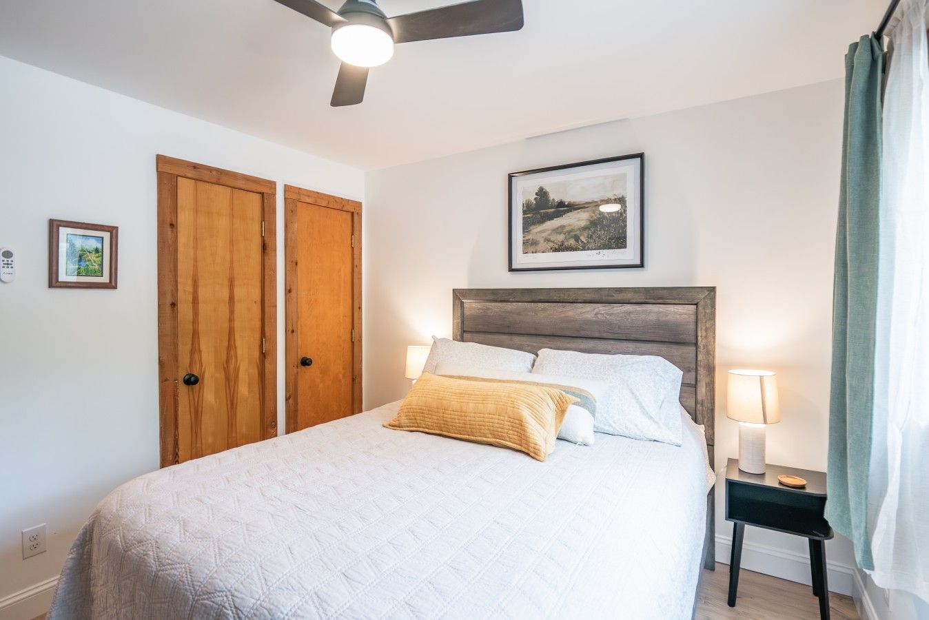 A bedroom with a bed, a bedside table with a lamp, a ceiling fan, and two wooden doors.