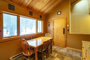 Un comedor con una mesa y sillas de madera, ventanas amplias con vistas a árboles y una puerta de madera.