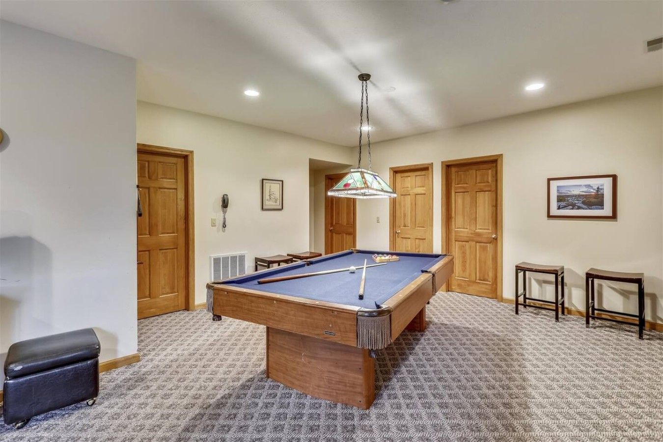 A pool table in a room with carpeted flooring, wooden doors, and a hanging light fixture.