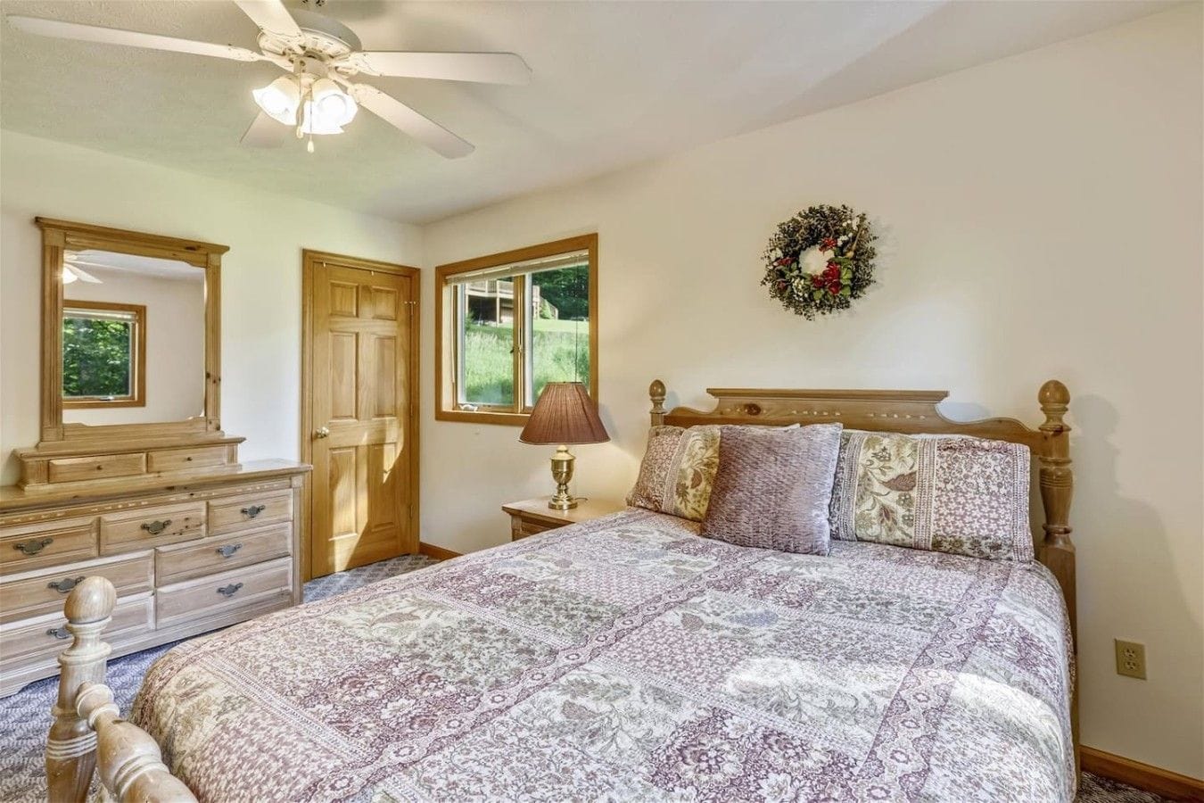 Une chambre avec un grand lit, une commode, une table de chevet avec une lampe, un ventilateur de plafond et une fenêtre donnant sur un paysage verdoyant.
