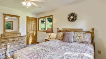 A bedroom with a large bed, a dresser, a nightstand with a lamp, a ceiling fan, and a window with a view of greenery.