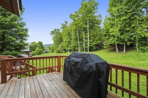 Une terrasse en bois dotée d’un barbecue, entourée d’une rambarde en bois et d’une végétation luxuriante.