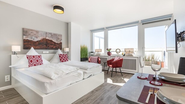 A modern hotel room with a large bed, a dining area with a table and chairs, a balcony with a view, and a mounted television. - Penthouse - Modern apartment with sea view, balcony, parking space, WLAN. Pets (Sylt)