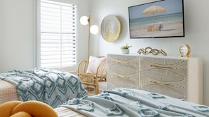 Une chambre à coucher comprenant un lit, une chaise, une commode, un téléviseur et un tableau sur le thème de la plage accroché au mur.