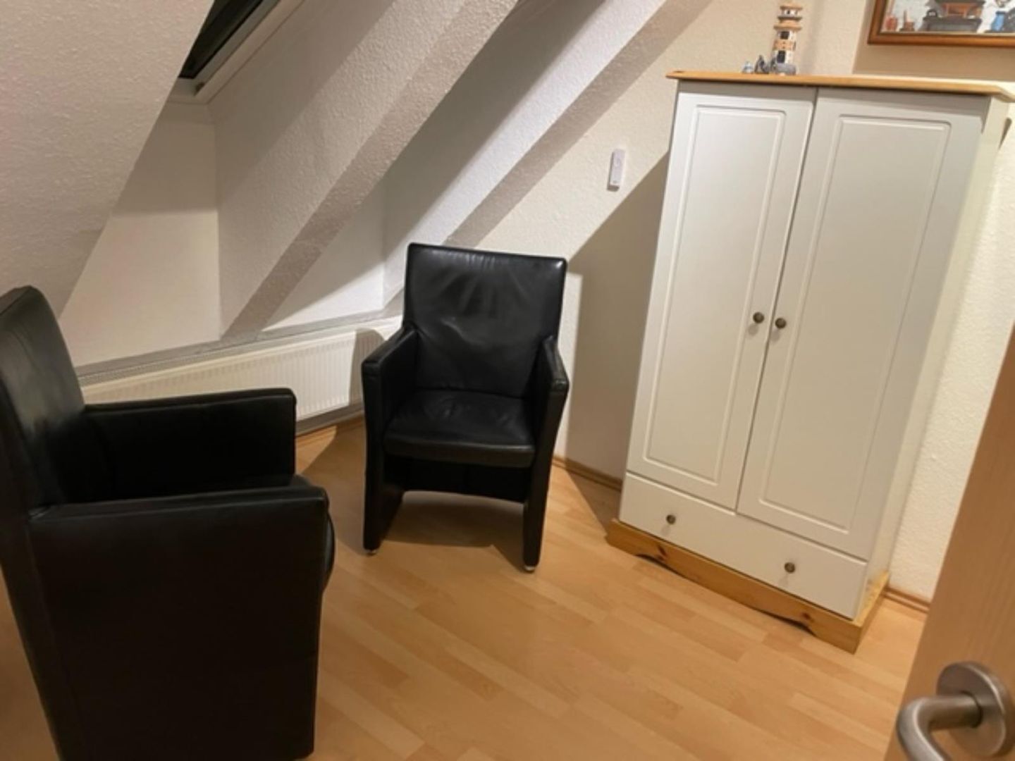 A room with a sloped ceiling, a black leather chair, a white cabinet, and a wooden floor.