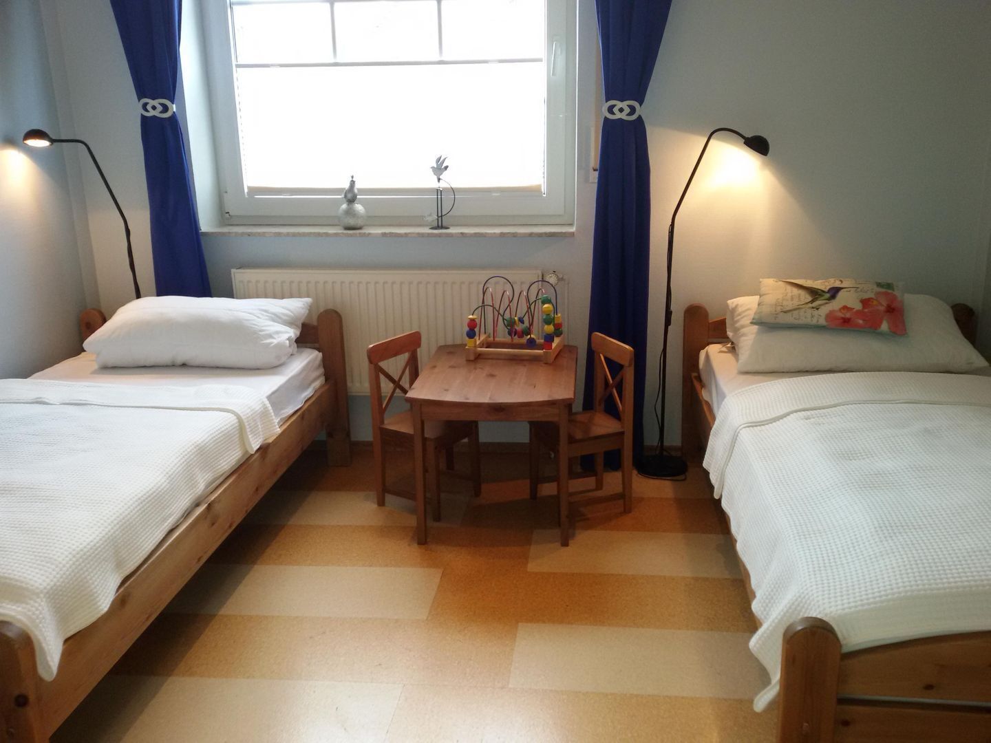 Quarto com duas camas de solteiro, uma mesa de madeira com cadeiras, um radiador e uma janela com cortinas.