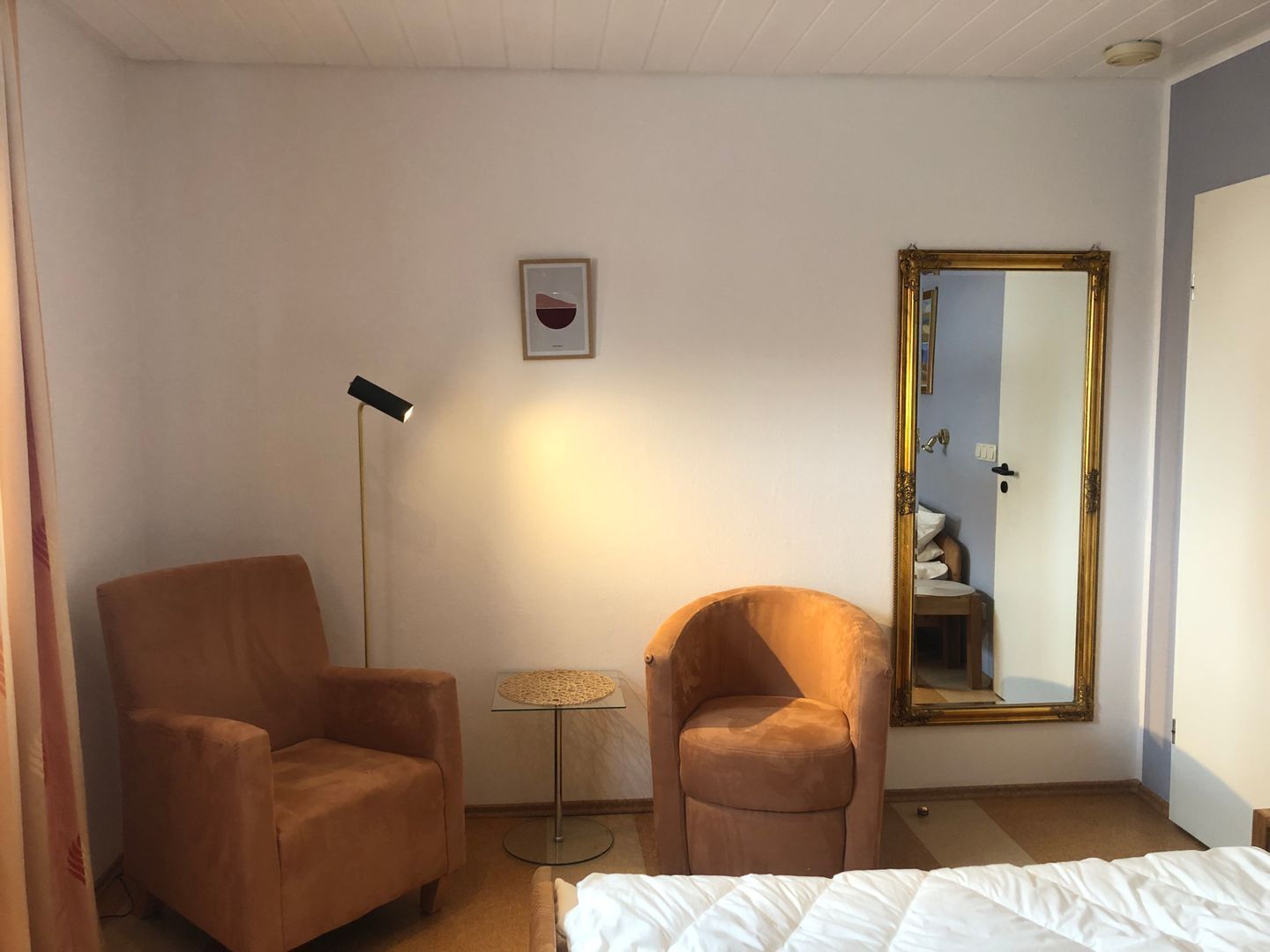 A hotel room with a bed, two armchairs, a floor lamp, a small table, and a large mirror.