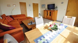 A living room with a sofa, a wooden coffee table, a dining table with a placemat, a television on a wooden cabinet, and two doors. - Vacation apartment Caspersweg 4a - Holiday apartment Fernblick Caspersweg 4a (Wittmund)