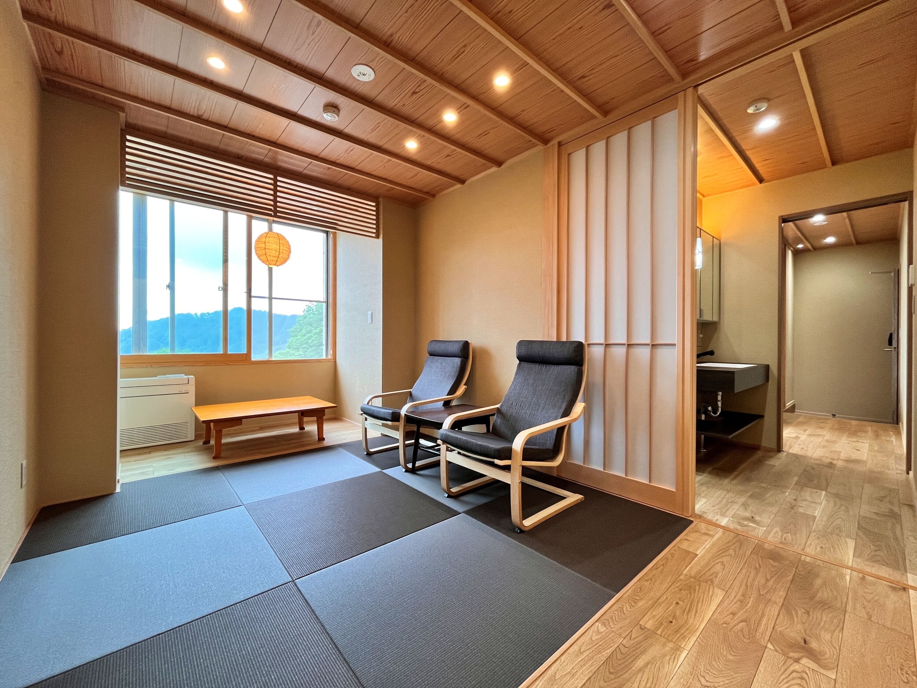 Una habitación con esteras de tatami, techo de madera, un banco, dos sillas, una ventana con persianas y vistas al exterior.