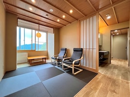 A room with tatami mats, a wooden ceiling, a bench, two chairs, a window with blinds, and a view of the outdoors.