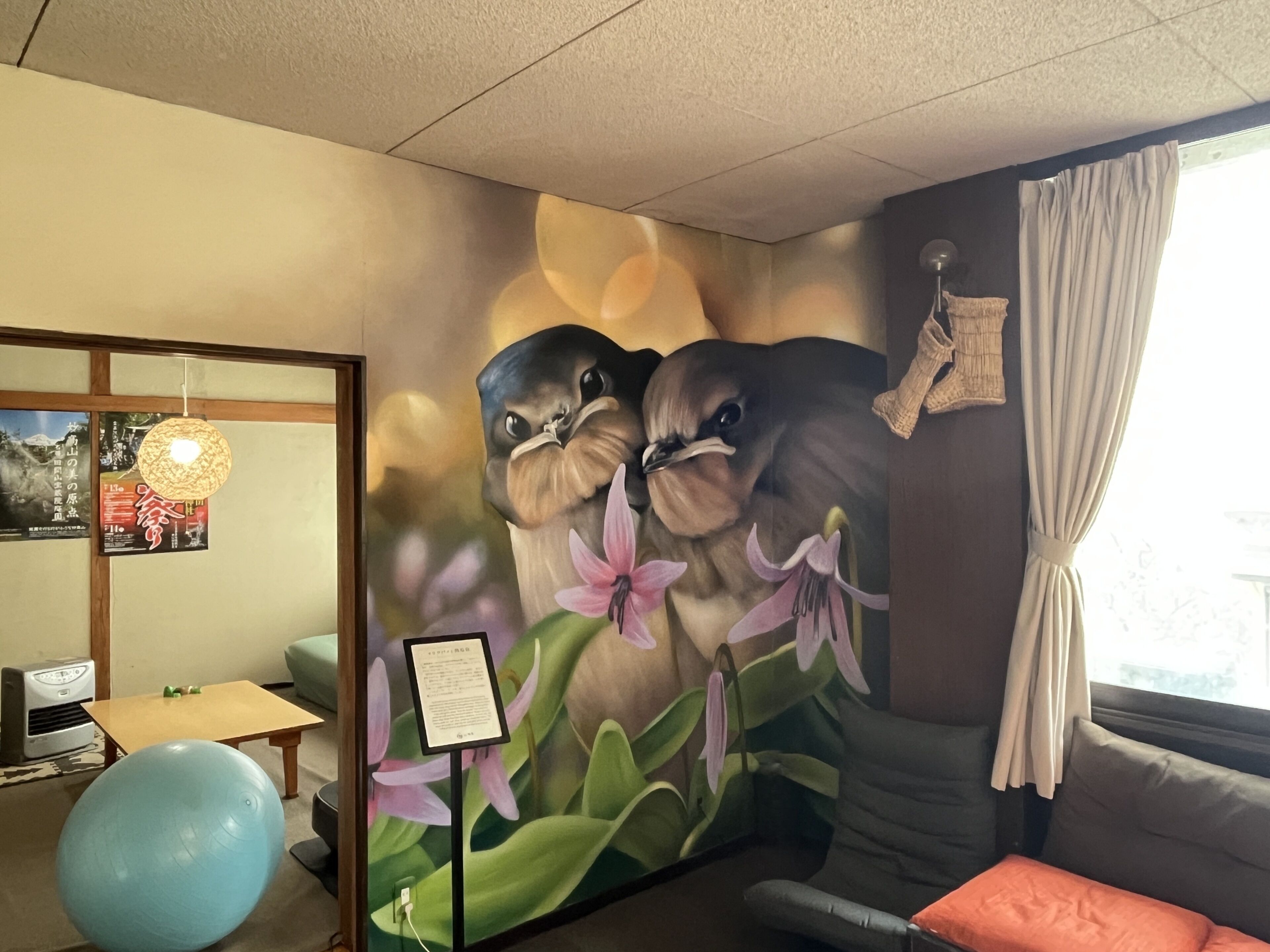 Une pièce décorée d’une fresque représentant des oiseaux et des fleurs, avec un ballon d’exercice bleu, une petite table, un canapé et une fenêtre avec des rideaux.