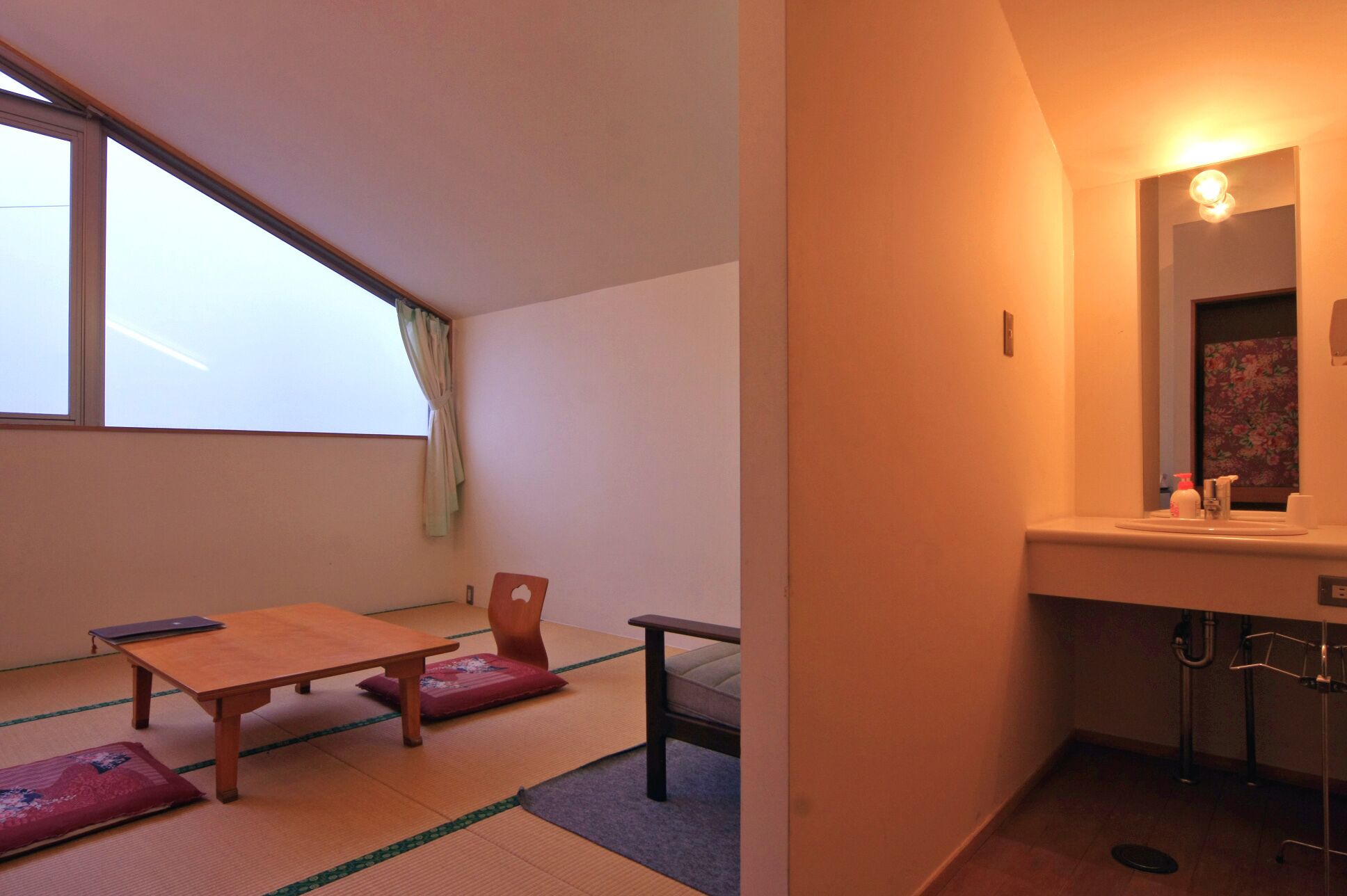 Une pièce avec des tatamis, une table en bois, une chaise, un petit tabouret et une salle de bain équipée d’un lavabo et d’un miroir.
