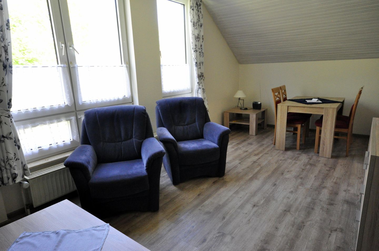 Ein Zimmer mit zwei Sesseln, einem kleinen Tisch mit Lampe sowie einem Esstisch mit Stühlen.
