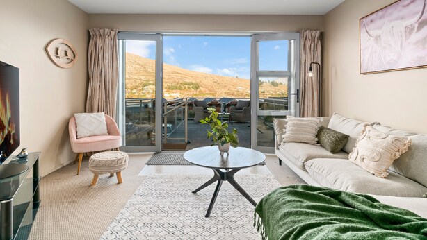 A modern living room with a sofa, a chair, a coffee table, and a TV. There is a sliding glass door leading to an outdoor area with a view of hills. - 2-BEDROOM REMARKABLES APARTMENT (Queenstown)