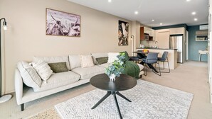 A modern living room with a sofa, coffee table, and artwork. A kitchen is visible in the background. - 2-BEDROOM REMARKABLES APARTMENT (Queenstown)