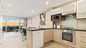 A modern kitchen with a dishwasher, stove, and microwave. There is a dining area with a table and chairs. - 2-BEDROOM REMARKABLES APARTMENT (Queenstown)