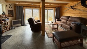 Een knus interieur met een leren bank, een fauteuil, een houten bankje, een kleine tafel en een groot raam met uitzicht op de natuur.