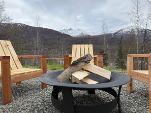 A circular fire pit with logs, surrounded by wooden chairs, set on gravel with a mountainous backdrop.