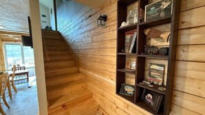 Een houten trap die leidt naar een kamer met een houten wand, een plank met diverse voorwerpen, en uitzicht op buiten.