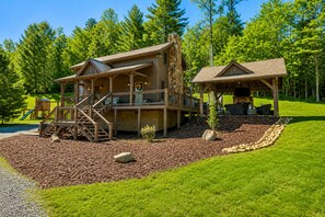 Eine zweistöckige Holzhütte mit einer Terrasse, einem überdachten Sitzbereich im Freien und einem Spielplatz.