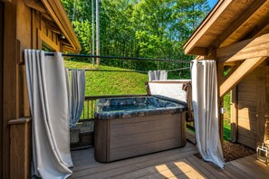 A wooden deck with a hot tub, curtains, and a view of a green landscape. - Farmhouse Cabin, Hot Tub, Fireplace Patio w/TV (Blue Ridge)