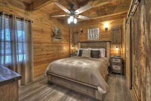 A wooden cabin bedroom with a bed, nightstands, a ceiling fan, and a 'HOME' sign on the wall. - Farmhouse Cabin, Hot Tub, Fireplace Patio w/TV (Blue Ridge)