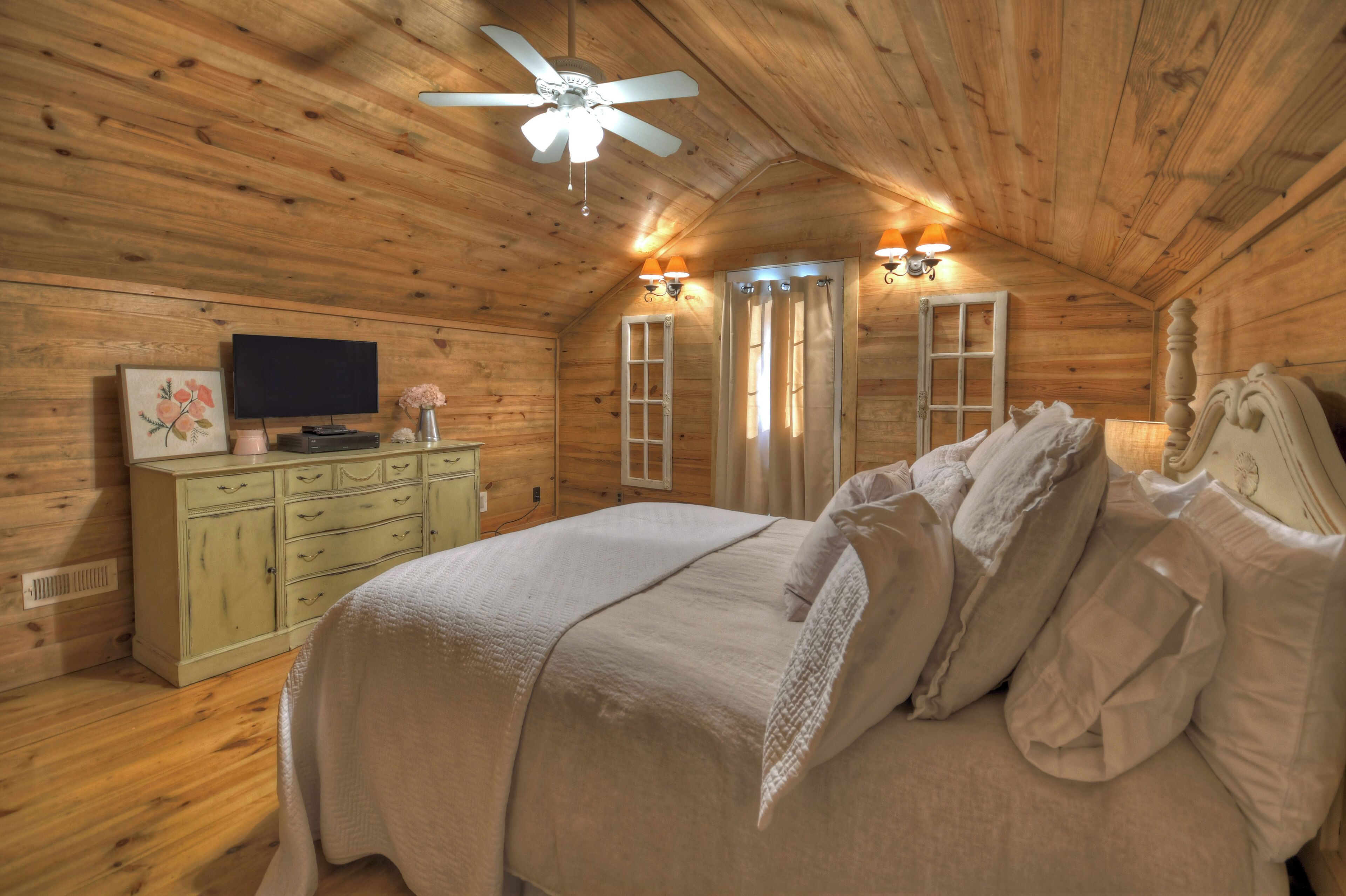 Een slaapkamer in een houten blokhut met een bed, een dressoir, een televisie en een plafondventilator.