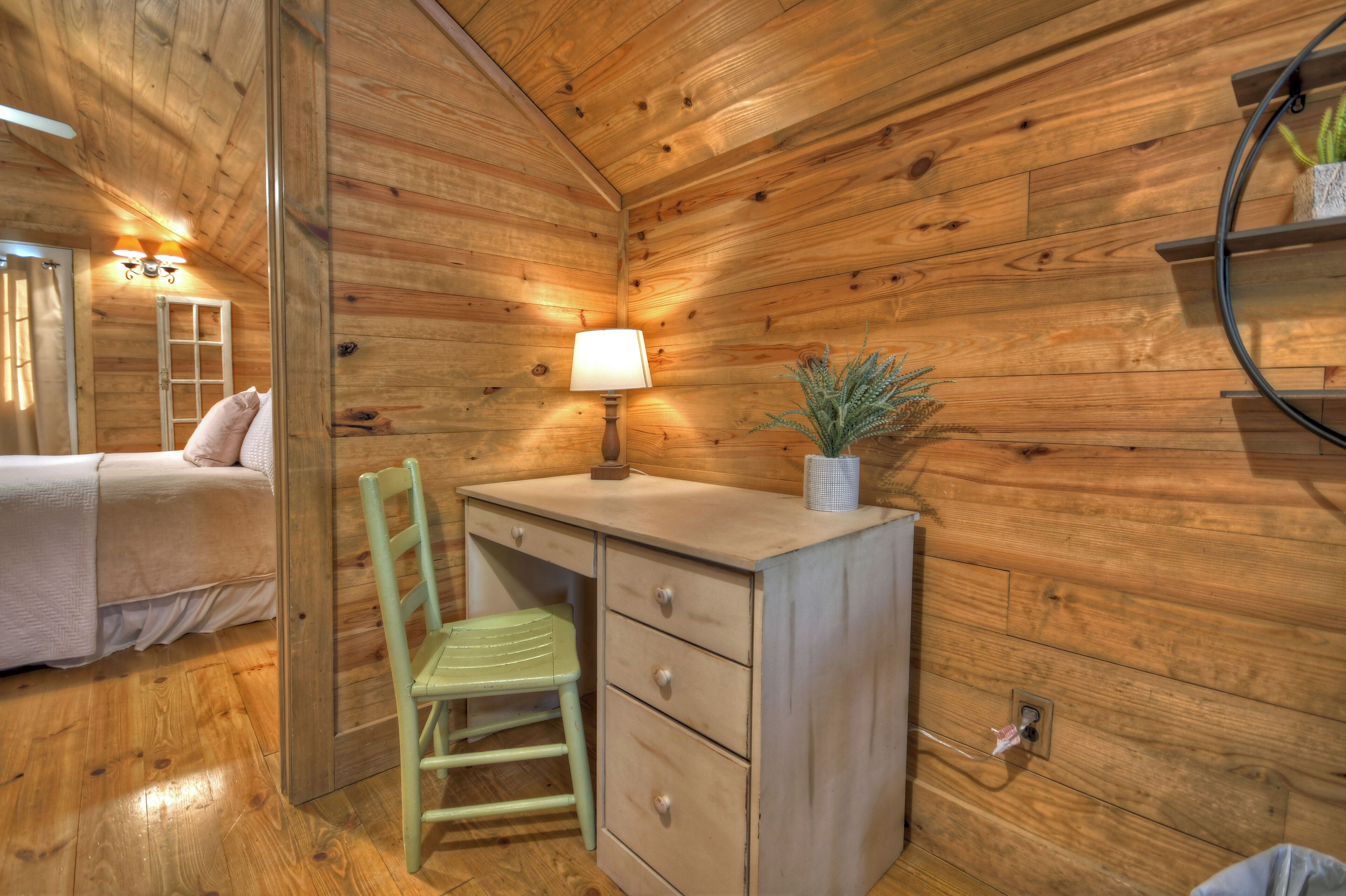 A wooden cabin room with a bed, a desk with a lamp, a chair, and a shelf with a plant.